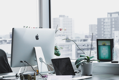 Apple laptop on a desk in an office against a backdrop of large windows looking out onto a large cityscape