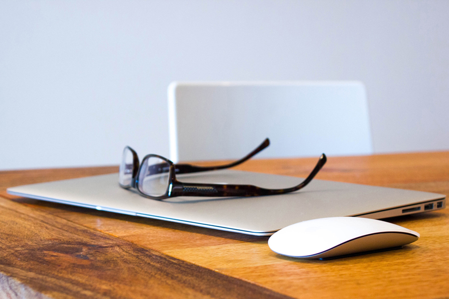 closed laptop, mouse, glasses resting on top of the laptop all on a desk